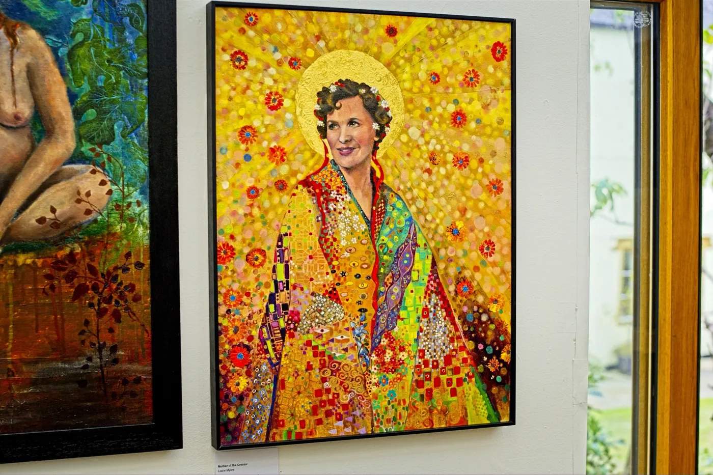 Two of Lizzie's paintings hanging in a sunlit gallery: a nude in a jungle and a golden mosaic portrait